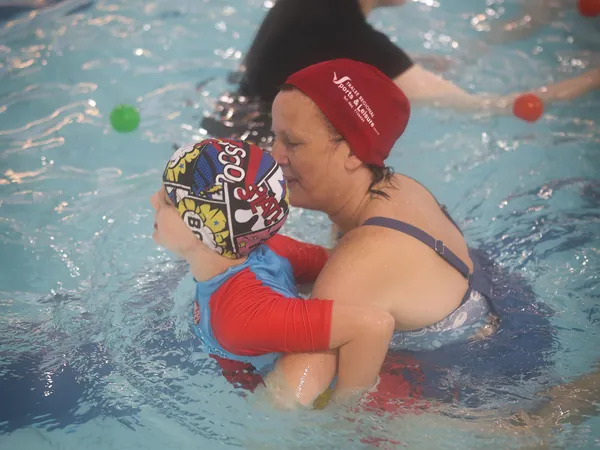 A parent holding a child wearing a red float during ball games in the pool