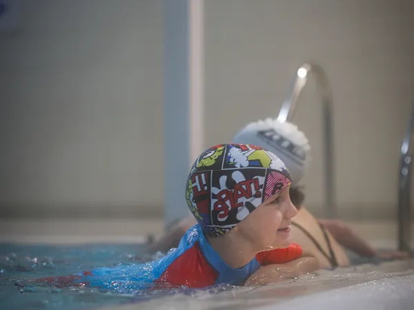 A child in a colourful comic-print swim cap swimming at the edge of the pool