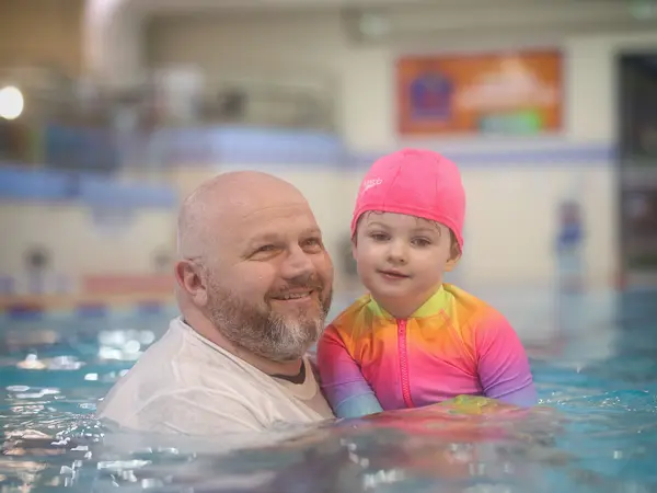 A father smiling with his young daughter in a colourful swimsuit in the pool