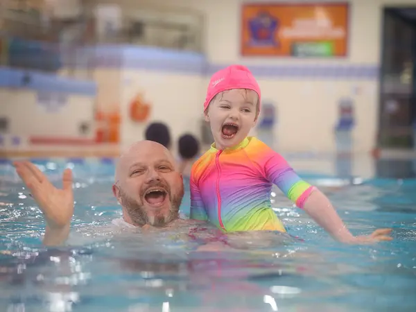 A father and his young daughter in a pink swim cap laughing together in the pool