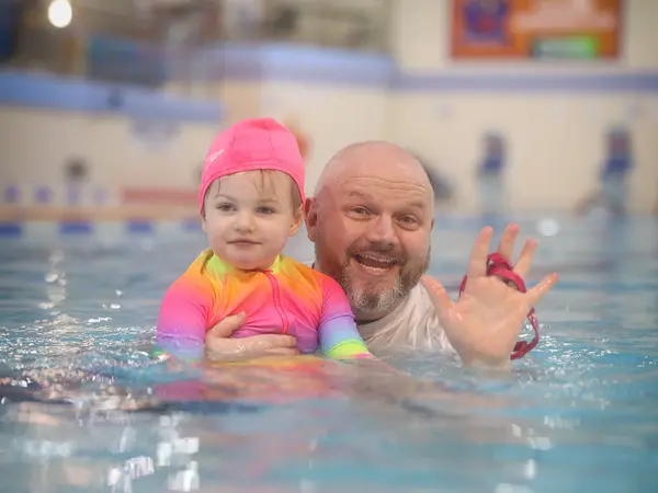 A toddler in a pink swim cap waving while held by her father in the pool