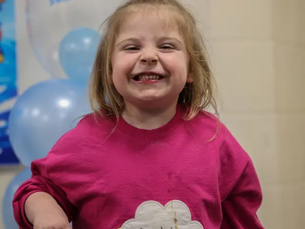 A girl in a pink jumper with a big smile in front of balloons
